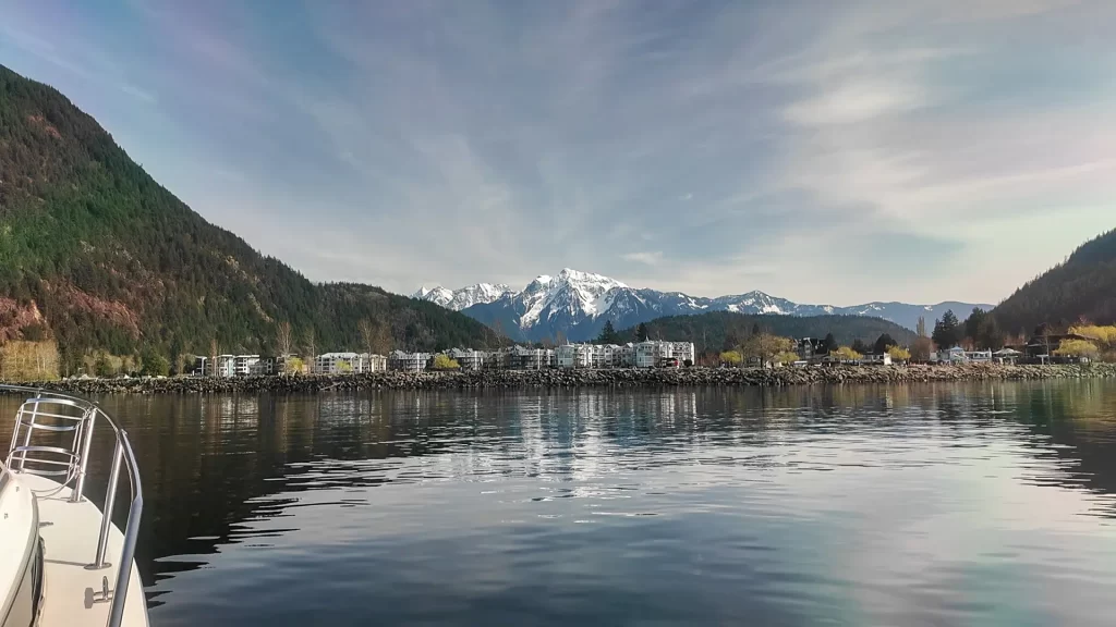A photo taken from a boat on Harrison Lake. The Township of Harrison Hot Springs is past the shore of the lake. The mountains of the Fraser Valley rise behind the town.